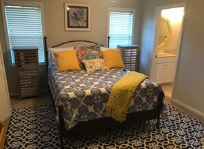 Une chambre à coucher comprenant un lit, deux tables de chevet, une commode, un miroir et une fenêtre avec des stores.