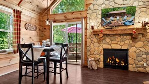 A wooden cabin interior with a stone fireplace, dining area, and a view of the outdoors.