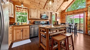 A wooden cabin with a kitchen featuring wooden cabinets, a stainless steel refrigerator, a microwave, and a dishwasher. There is a large wooden dining table with two stools.