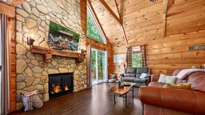A log cabin interior with a stone fireplace, wooden walls, a leather sofa, and a large window.