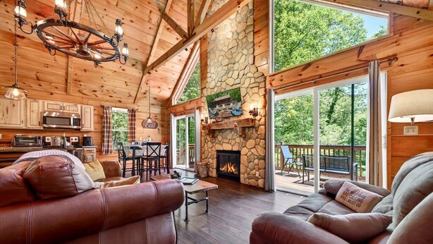 A log cabin interior with a stone fireplace, leather sofas, a dining area, and a large sliding glass door to the outdoors.