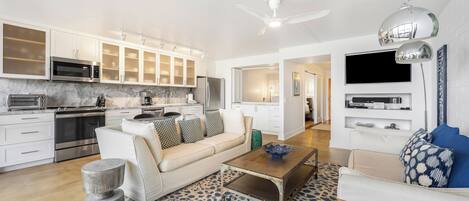 A modern kitchen with white cabinets, a microwave, and a stove. There is a living area with a sofa, coffee table, and a TV.