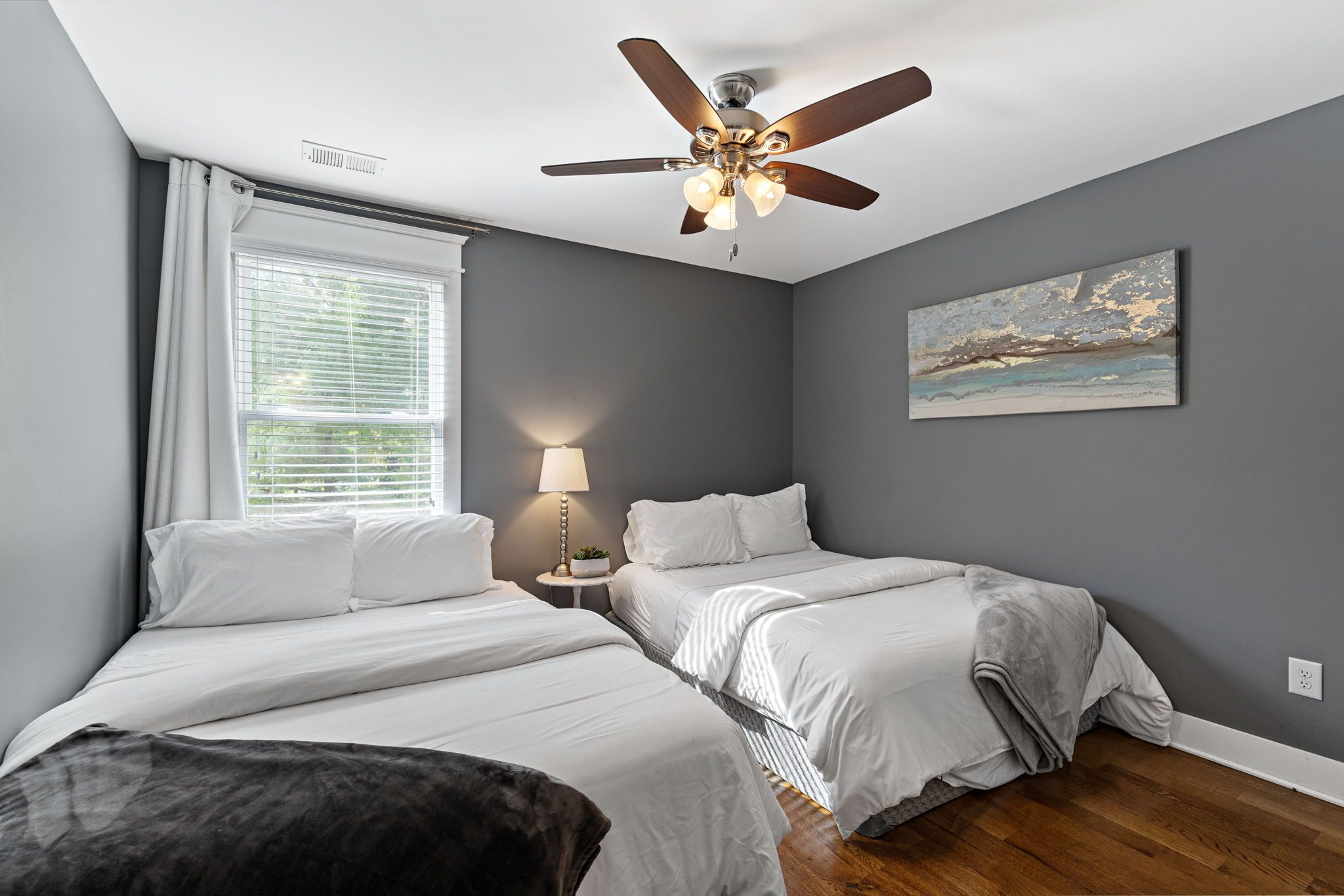 Une chambre avec deux lits, un ventilateur de plafond, une fenêtre avec des stores, une lampe et un tableau au mur.