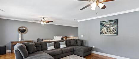 Un salon moderne avec un canapé d’angle, deux ventilateurs de plafond, un plancher en bois et une œuvre d’art encadrée accrochée au mur.