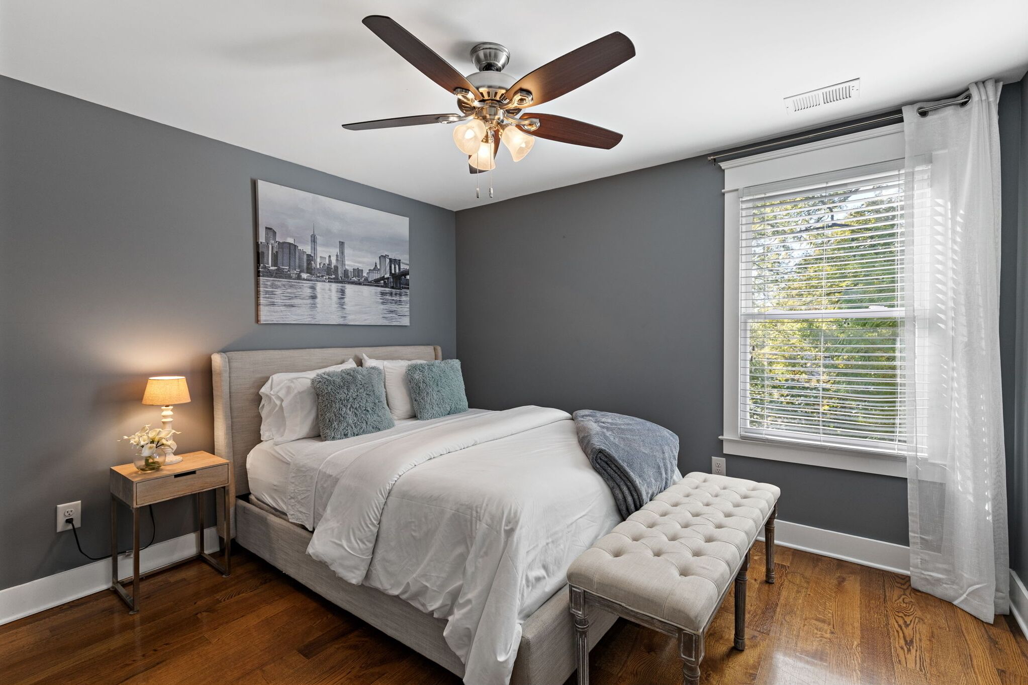 A bedroom with a bed, bedside table, lamp, and a cityscape painting on the wall.