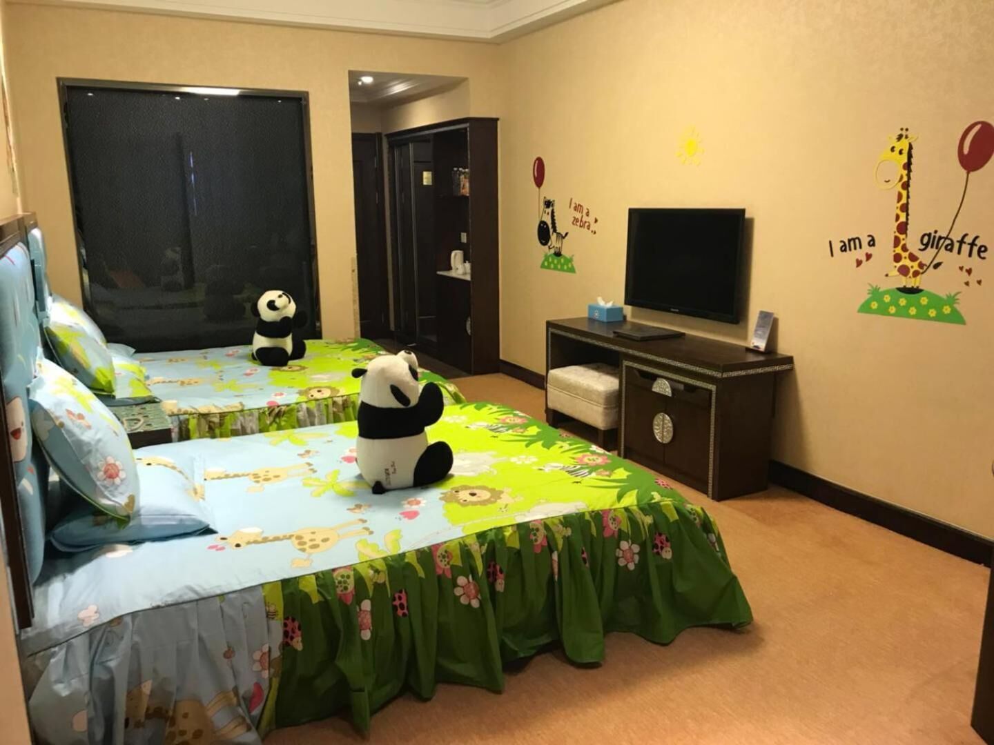 A hotel room with two beds, each with a panda plush toy and floral bedspreads. There is a TV on a wooden stand and a wall with animal-themed decorations.