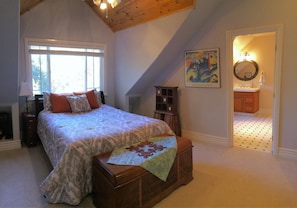 A bedroom with a large bed, a wooden chest, a window, a painting, and a bathroom visible through an open door.