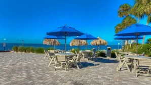 Un patio frente a la playa con mesas y sillas, sombrillas azules y sombrillas de paja.