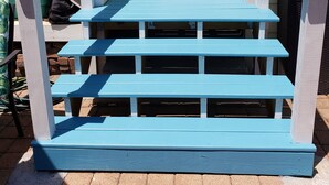 Unos escalones de madera azul conducen a una puerta blanca con un ventanal, frente a una casa amarilla.
