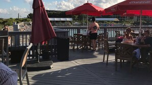 An outdoor dining area with wooden tables and chairs, red umbrellas, and a view of the water.