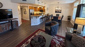 A spacious living room with a large TV, comfortable seating, a kitchen in the background, and a dining area with a table and chairs.