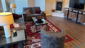 A living room with a sofa, coffee table, rug, ottoman, TV stand, fireplace, and a clock on the wall.