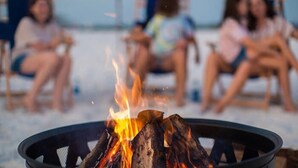 Un pozo de fuego con leña ardiendo en primer plano, rodeado de sillas de playa y gente sentada al fondo.