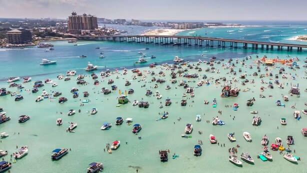 Un puerto deportivo con numerosas embarcaciones amarradas, un muelle que se adentra en el agua y una vista distante de una zona turística.