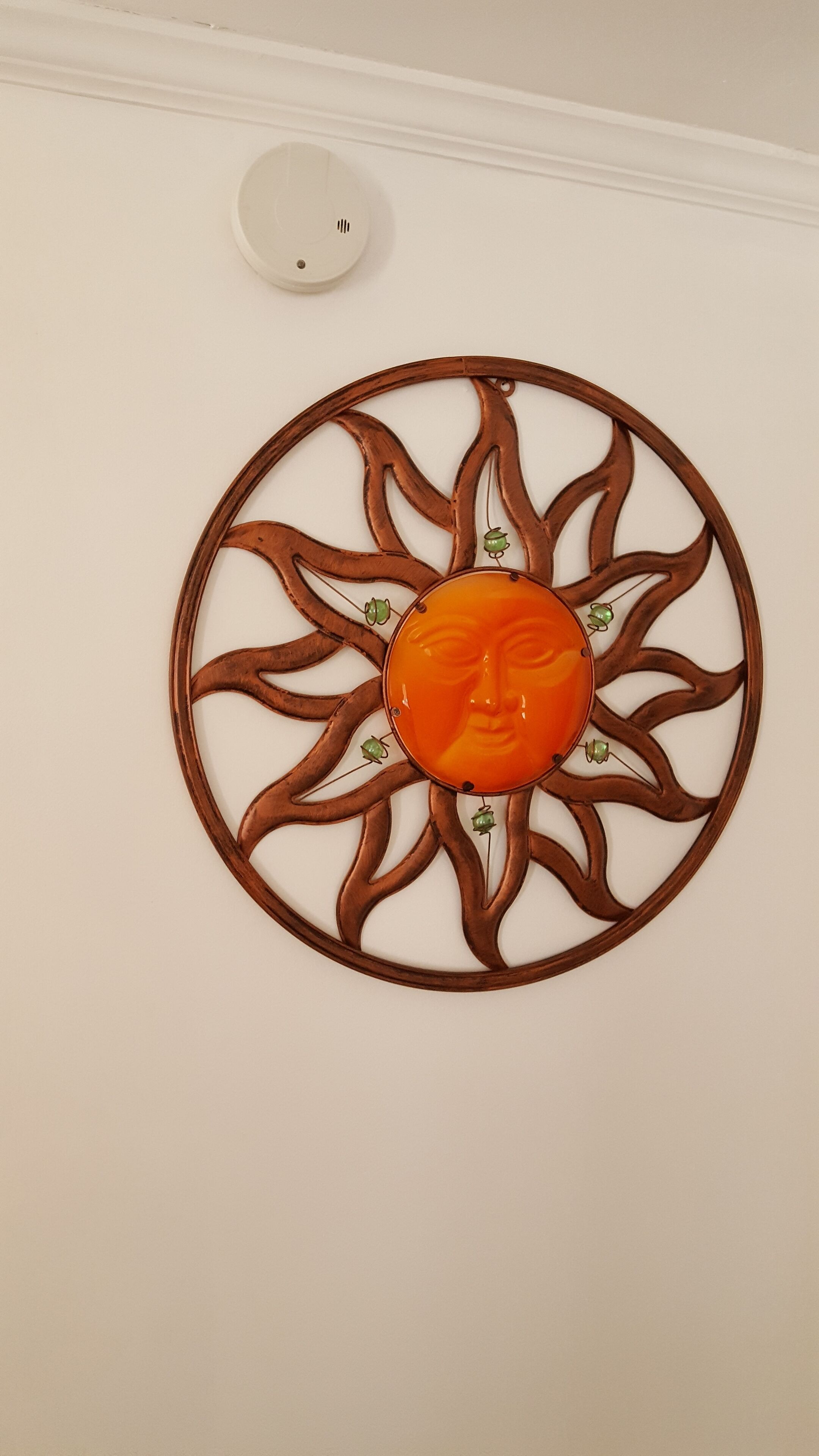 Un élément décoratif mural, fixé au mur, avec un motif central orange en forme de visage, entouré d’un décor évoquant un soleil.
