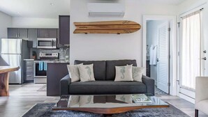Una sala de estar moderna con un sofá gris, un cuadro de un tabla de surf de madera, una mesa de centro de vidrio y una cocina al fondo.