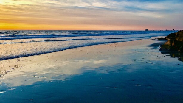 A beach with waves, a pier in the distance, and a clear sky.