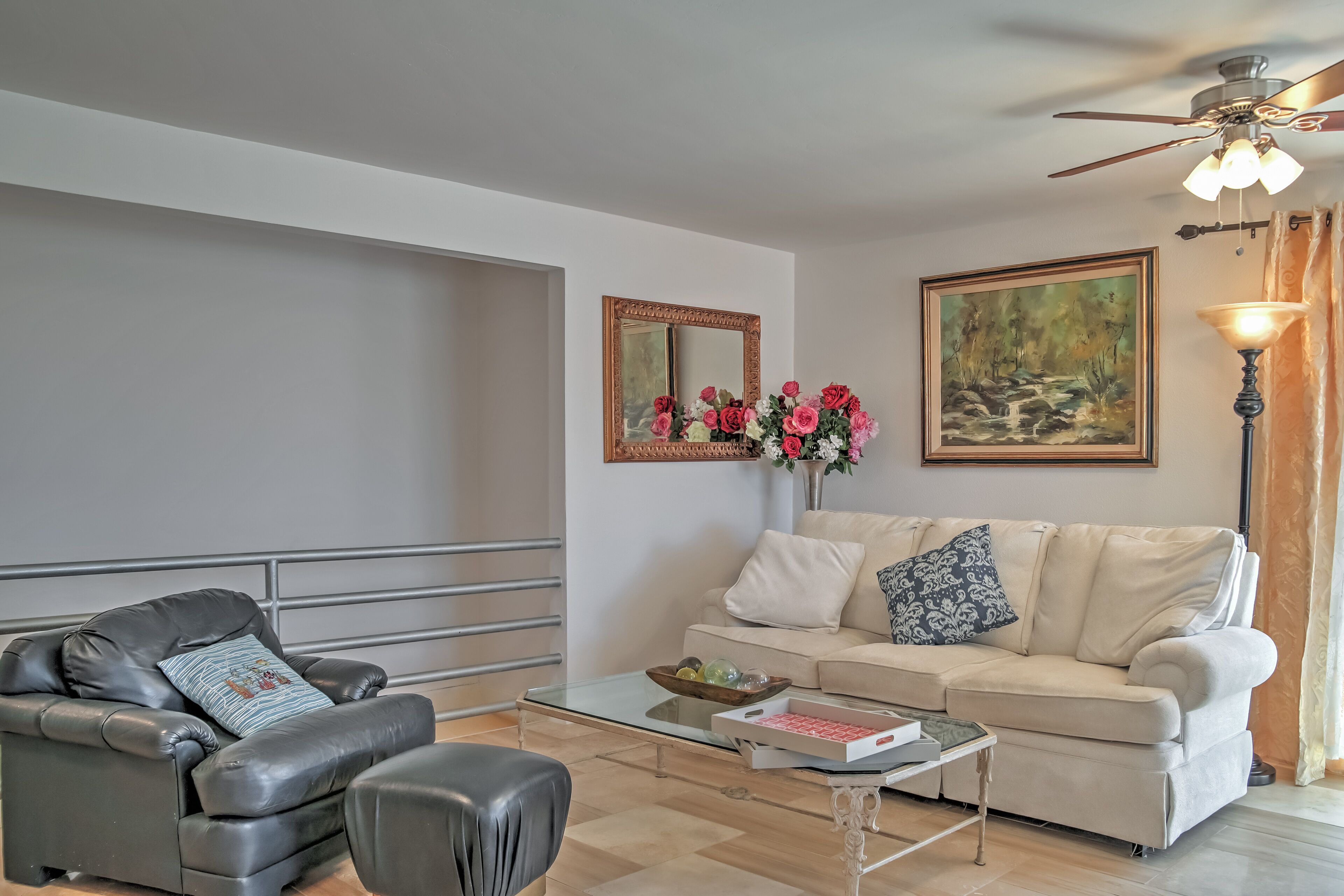 A living room with a black leather armchair, a beige sofa, a glass coffee table, a framed mirror, a painting, and a floor lamp.