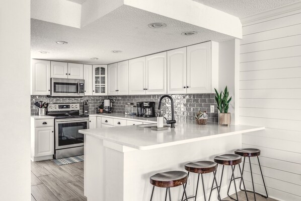 A modern kitchen with white cabinets, a central island with stools, a microwave, stove, and a coffee maker.