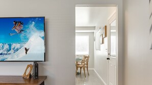 Un pasillo con una televisión mostrando a un esquiador, una mesa de madera con libros y un objeto decorativo, y un comedor con sillas y una mesa.