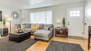 Una sala de estar con un sofá gris, un reposapiés negro, una mesita de madera y una puerta blanca.