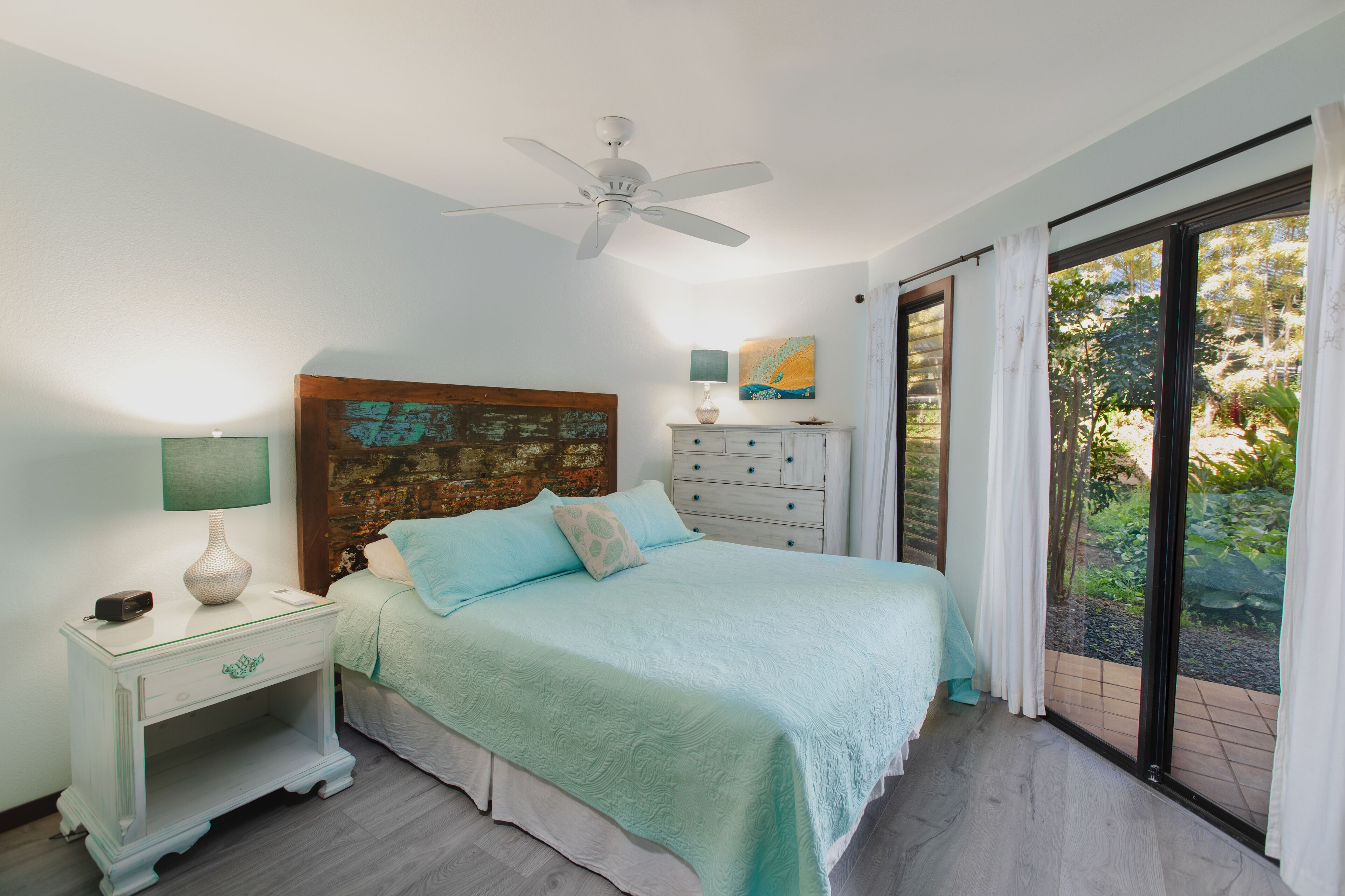 A bedroom with a bed, nightstand, dresser, ceiling fan, and large sliding glass door leading to a garden.