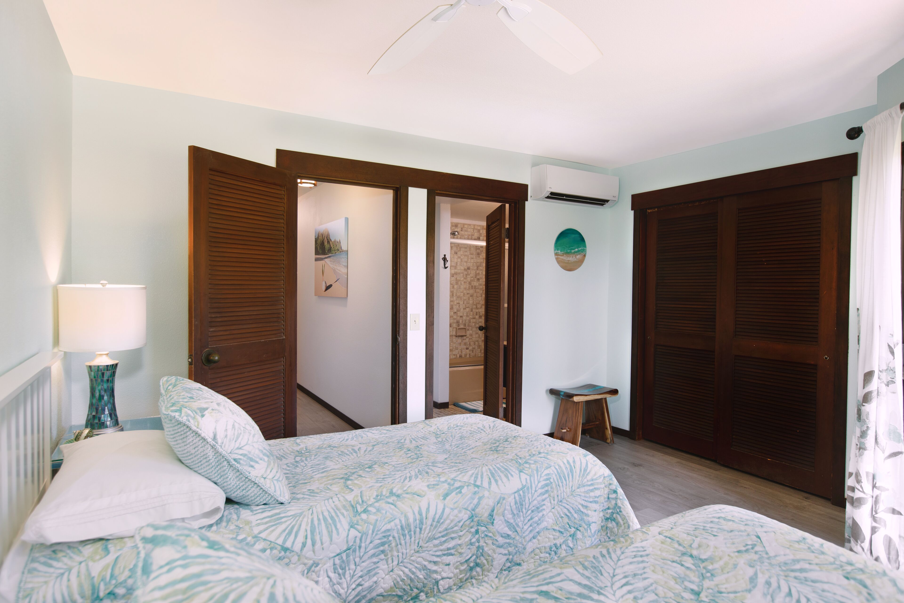 A bedroom with a bed, a nightstand, a lamp, a wall-mounted air conditioner, and a door leading to another room.
