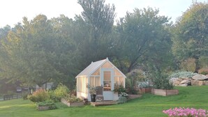 Una pequeña estructura blanca con techo a dos aguas y puerta de vidrio, rodeada por un jardín cuidado con diversas plantas y un gran árbol al fondo.