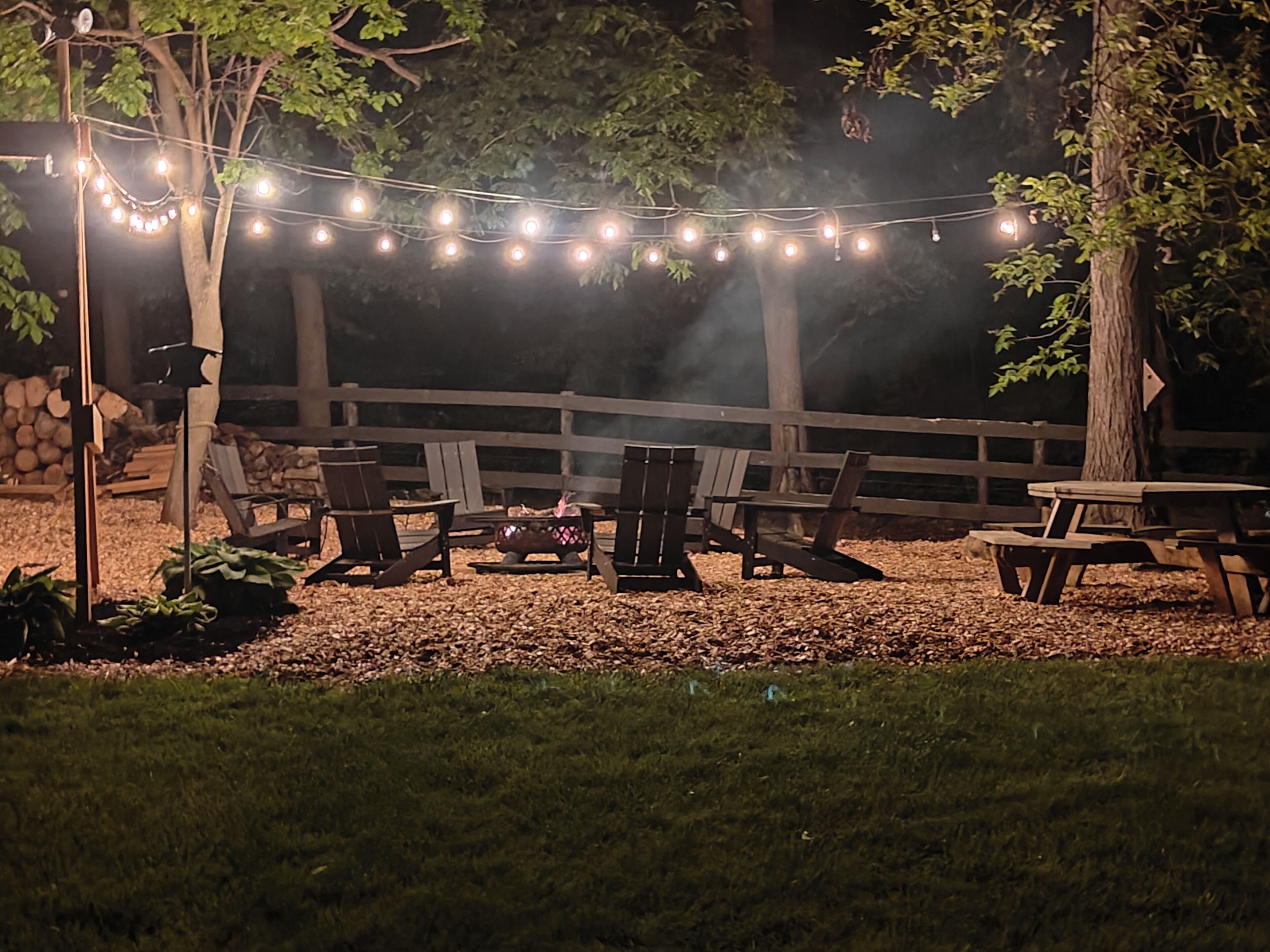 An outdoor seating area with string lights, wooden chairs, a fire pit, and a wooden fence.