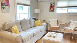 A living room with a beige sofa, two white armchairs, a small table, and a white ottoman. There are two windows with blinds, a wall-mounted air conditioner, and two paintings. - Fort Grounds Cottage Across from City Beach, Park & Downtown! (Coeur d'Alene)