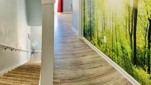 Un pasillo con piso de madera, un pilar blanco y un mural de un bosque en la pared derecha.