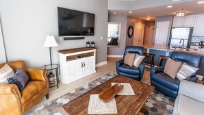 A modern living room with a TV, leather sofas, a wooden coffee table, and a kitchen in the background. - Lighthouse 910 - Perfect Spot in the Middle of the Building (Gulf Shores)