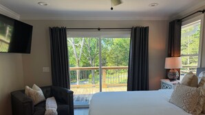 A bedroom with a large bed, a dark leather chair, a small table with a lamp, and a view of a wooden deck and trees.