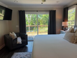 A bedroom with a large bed, a dark leather chair, a small table with a lamp, and a view of a wooden deck and trees.