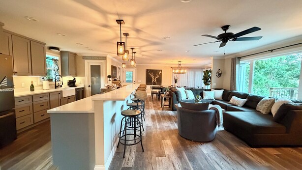 A modern kitchen with a central island, bar stools, a dining area with a piano, and a living room with a sofa and a ceiling fan.
