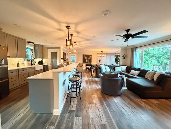 A modern kitchen with a central island, bar stools, a dining area with a piano, and a living room with a sofa and a ceiling fan.