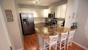 Una cocina moderna con armarios blancos, encimera de granito, refrigerador de acero inoxidable, microondas y cafetera.