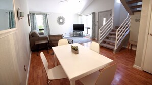 Amplia sala de estar con una mesa de comedor, sofá y televisor. Una escalera con barandillas blancas conduce a otro nivel.
