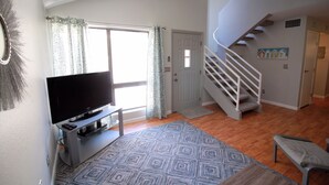 Una sala de estar con televisor de pantalla plana, alfombra estampada, una escalera y una puerta con un pequeño ventanal.