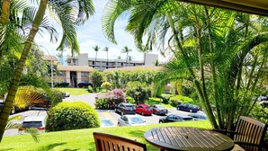 Une vue depuis un balcon, avec une table et des chaises en bois, donnant sur un parking où sont garées plusieurs voitures, le tout entouré d’une végétation luxuriante et de palmiers.