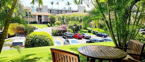 A view from a balcony with a wooden table and chairs, overlooking a parking lot with several cars, surrounded by lush greenery and palm trees.