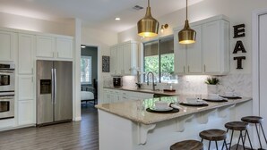 A modern kitchen with white cabinets, a stainless steel refrigerator, a granite countertop, and bar stools. - Walk to Coachella! 7BR~Sleeps 14~Pool~Game Room! (Indio)