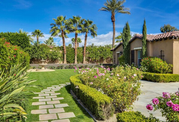 A well-maintained garden with a stone pathway, palm trees, and flowering bushes.