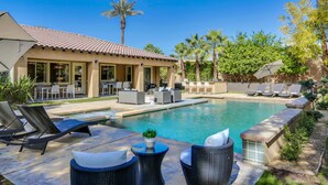 A pool area with lounge chairs and a patio table, surrounded by a house with a tiled roof and a covered outdoor seating area. - Walk to Coachella! 7BR~Sleeps 14~Pool~Game Room! (Indio)