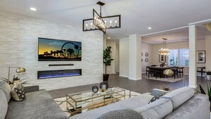 A modern living room with a large flat-screen TV, a fireplace, and a dining area with a chandelier. - Walk to Coachella! 7BR~Sleeps 14~Pool~Game Room! (Indio)