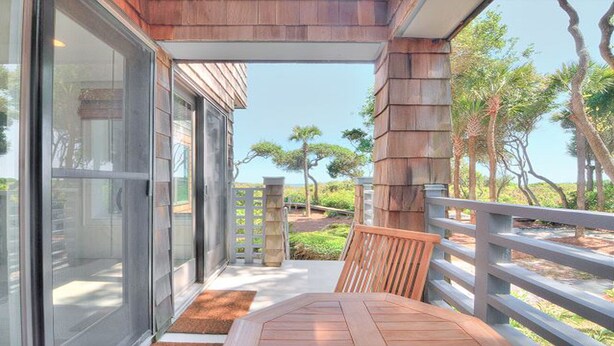 A balcony with a wooden table and chairs, a glass door leading to a patio, and a view of a landscaped area with trees. - Spectacular 1 Bedroom, Oceanfront Villa! BRAND NEW RENOVATION!! (Kiawah Island)