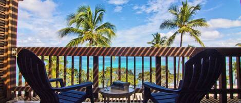 A covered outdoor area with two chairs and a table, overlooking a beach with palm trees.