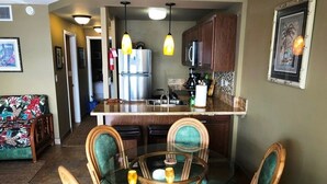 Un comedor con una mesa de vidrio rodeada de sillas de mimbre, una cocina al fondo y un cuadro en la pared.