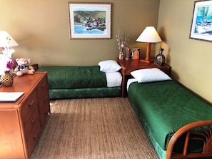Habitación de hotel con dos camas verdes, un aparador de madera, un escritorio con lámpara y dos cuadros en la pared.