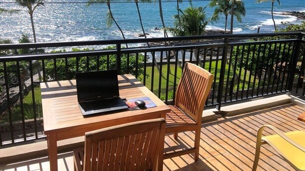 Una terraza de madera con una mesa y una silla, con vistas a una playa con palmeras y el océano.